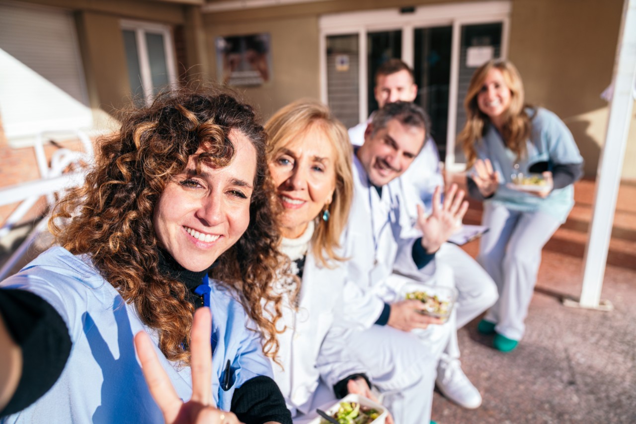 Outdoor gathering of medical staff, with one nurse standing and talking, while others sit, enjoying healthy snacks and engaging discussions