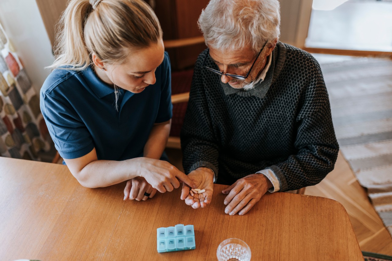 Verzorgende helpt oudere man met medicijnen aan tafel