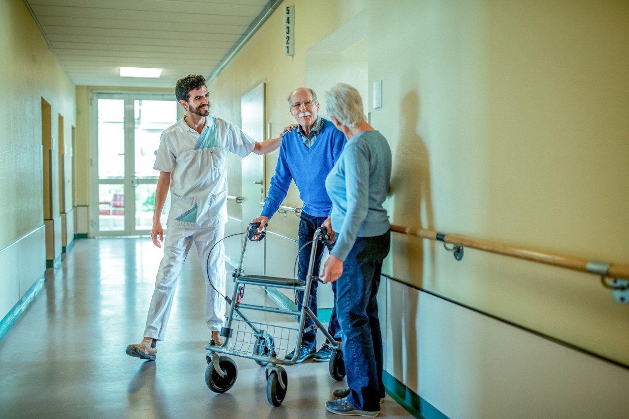 Ouder echtpaar waarvan man met rollator samen met verpleegkundige op gang
